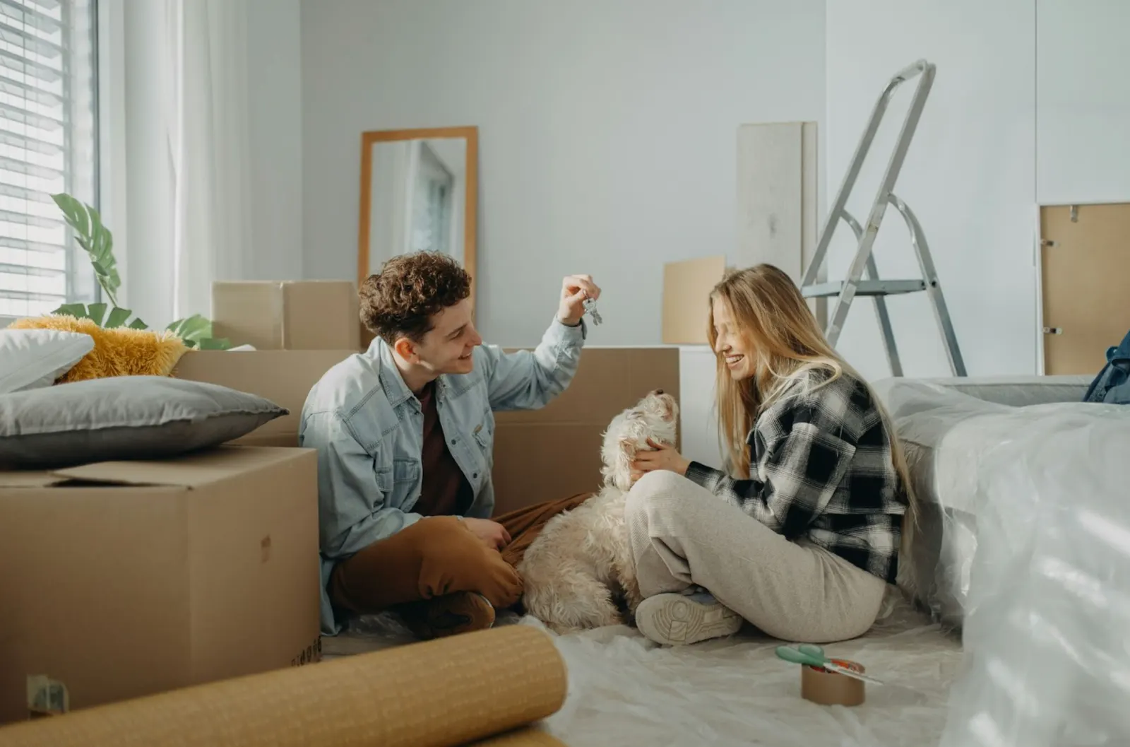 A young couple sitting on the floor surrounded by moving boxes, smiling and holding up a house key, with a small dog beside them.