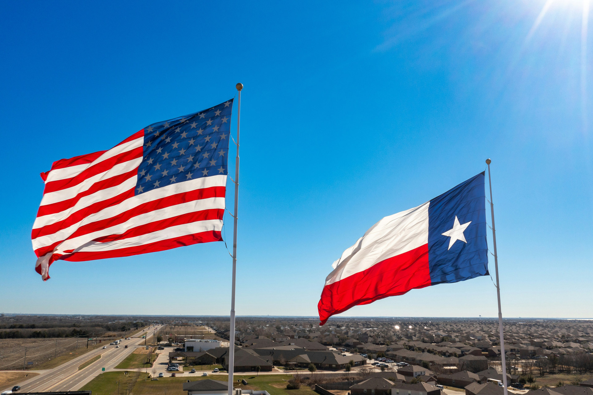 
The Texas and American flags wave proudly under a clear blue sky, symbolizing unity and pride over a sprawling suburban landscape.