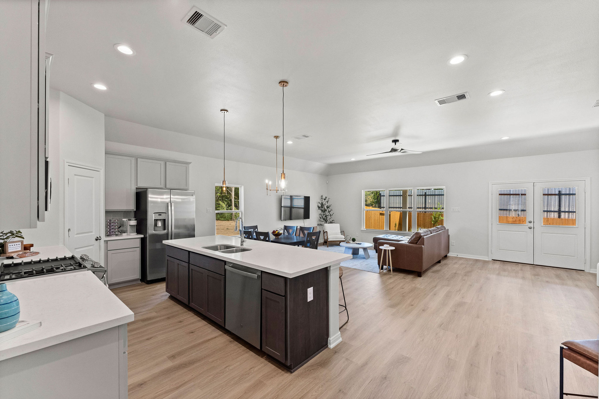 Modern open-plan kitchen and living room with a large island, stainless steel appliances, wooden flooring, and a brown sofa by a window.