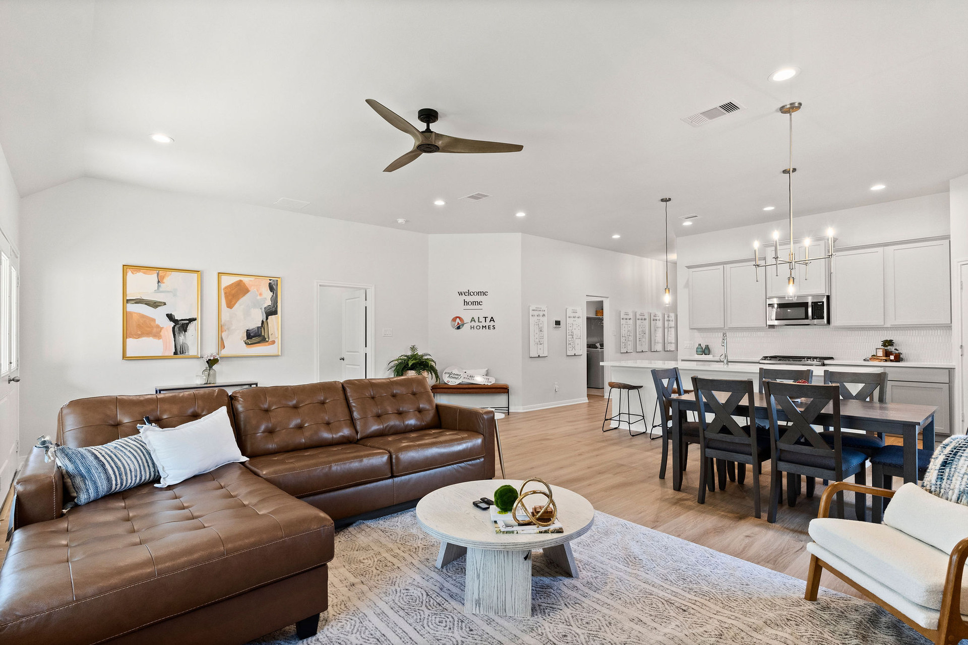 Modern living room and kitchen with a brown leather sofa, abstract art, and a round coffee table. "Welcome home" text on wall. Cozy ambiance. Lexington Heights in Willis, Texas
